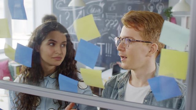 Chest-up shot of creative Caucasian guy and girl discussing mind map for creative new project and arranging colorful sticky notes on glass wall in office, and colleagues working in background