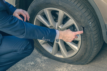 A businessman in a blue suit crouching near his car and checks the degree of damage to a punctured wheel. Hole in the tire. Concept - obrazy, fototapety, plakaty