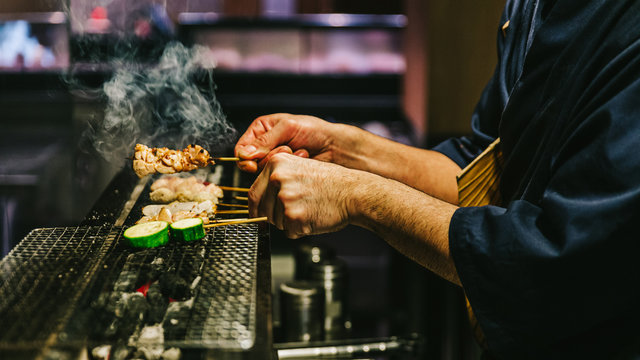 Hands of Japanese Yakitori Chef grilling chicken marinated with ginger, garlic and soy sauce and cucumber with a lot of smoke.