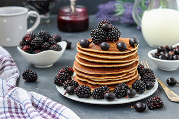 Pancakes with maple syrup, blackberries and currants are stacked on a plate, in the background berries and a jug of milk, horizontal orientation