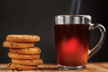 A pile of oatmeal cookies with chocolate chips and a mug of fragrant black hot tea in on a bamboo substrate, on a dark background. Handmade cookies for a healthy breakfast