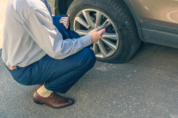 A businessman in suit crouching near his car with  punctured wheel. Man calling police using mobile...