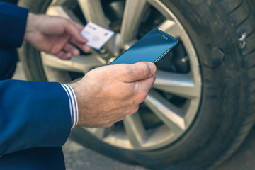 A businessman in suit crouching near his car with  punctured wheel. Man calling  tow truck service using mobile phone. Technicial help on the road. concept - obrazy, fototapety, plakaty