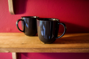 Two black coffee mugs are sitting on a wooden kitchen shelf