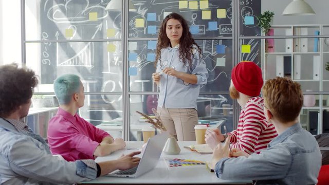 Medium shot of group of young Caucasian woman standing in office in front of glass panel with notes and teaching masterclass for group of creative people, who are listening and asking questions