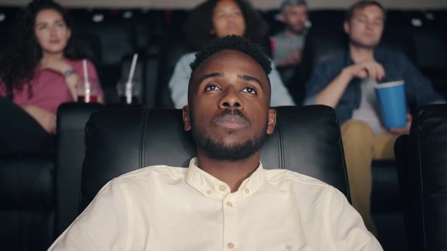 Sleepy African American student bearded guy is watching movie in cinema closing eyes winking relaxing in comfortable seat. Youth and relaxation concept.