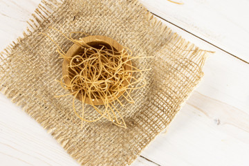 Lot of pieces of thin raw pasta noodles in wooden bowl on jute cloth flatlay on white wood
