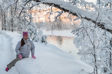 Fototapeta na wymiar Young Caucasian woman sitting in snow in winter forest, near open water river