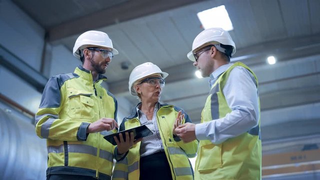 Three Heavy Industry Engineers Stand in Pipe Manufacturing Factory, Use Digital Tablet Computer, Have Discussion. Design and Construction of Large Oil, Gas and Fuels Transport Pipeline. Low Angle