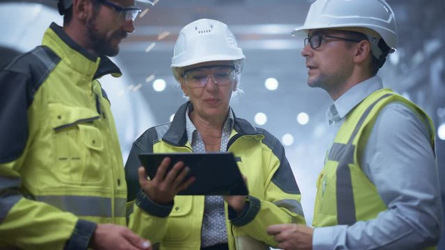 Three Heavy Industry Engineers Stand in Pipe Manufacturing Factory, Use Digital Tablet Computer, Have Discussion. Large Pipe Assembled. Design and Construction of Oil, Gas and Fuels Transport Pipeline