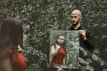young guy holds a mirror in the forest in the rain. a girl is reflected in a mirror. concept of relationship, love, feelings, soul.