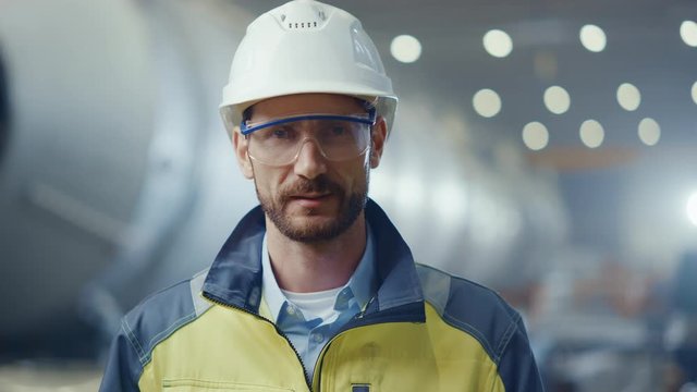 Portrait of Professional Heavy Industry Engineer / Worker Wearing Safety Uniform, Goggles and Hard Hat Smiling. In the Background Unfocused Large Industrial Factory where Welding Sparks Flying