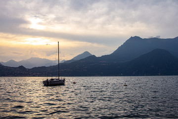 Fototapeta premium Night View to sailboat anchored in harbor the village of Varenna on Lake Como. Scenic landscape sunset on background alps mountains and water. Milan Province of Lecco. Italian Lombardy, Italy, Europe