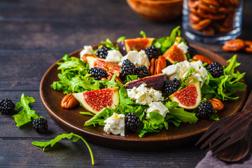 Salad with figs, feta cheese and blackberries in a wooden plate on dark background.