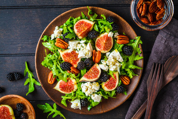 Salad with figs, feta cheese and blackberries in a wooden plate on dark background, top view.
