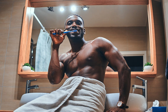 Attractive young man in towel is brushing his teeth in the bathroom while sitting on the table.