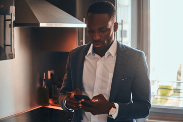 Young african man in white shirt is chatting by mobile phone at the kitchen.