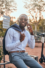 Cheerful black man in sunglasses is chilling in the park while talking by mobile phone.