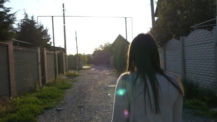 Slow motion, a girl with long hair is walking along the street between houses at sunset