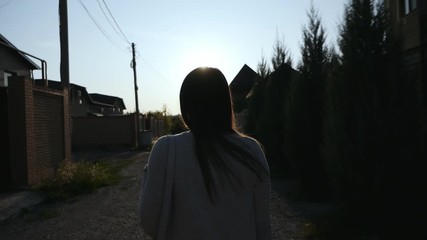 Shooting from the back, waters from the back, a girl with long hair is walking along the street between houses at sunset