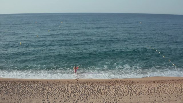 Drone shot of slim female running with color inflatable mattress along sandy beach to blue water of Mediterranean sea. Rear view of woman in lilac bikini swimming on pool float during summer leisure