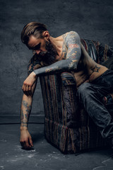 Portrait of tattooed sad man in despair, which is sitting on armchair at dark stash.
