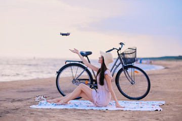 Obraz premium Pretty happy woman is trying to catch a drone while chilling on the beach next to her bike.