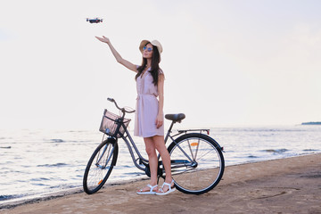 Fototapeta premium Pretty happy woman is trying to catch a drone while chilling on the beach next to her bike.