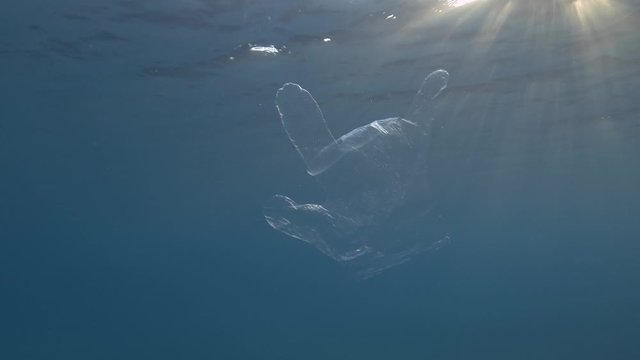  Plastic pollution, Discarded plastic glove slowly drifting under surface of blue water in the sunset light. Transparent plastic glove floats in Mediterranean Sea, Europe. Slow motion