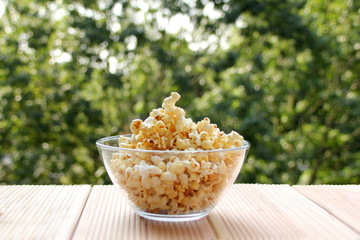 glass cup with popcorn on a light wooden table against a green summer garden, close-up, copy space