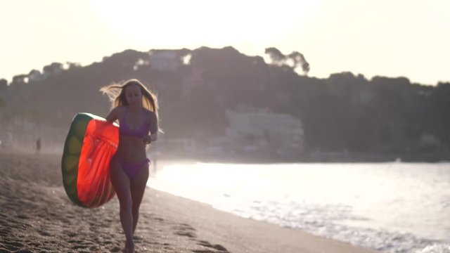 Beautiful female with long blonde hair walking on sandy beach at sunrise holding air lilo ball. Slim woman in lilac bikini with inflatable mattress enjoying summer leisure on sea coast of resort town