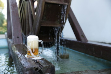 Fototapeta na wymiar Glass of beer with foam on a natural wooden board on a background of a wooden mill wheel with falling water. Pattern, banner, background.