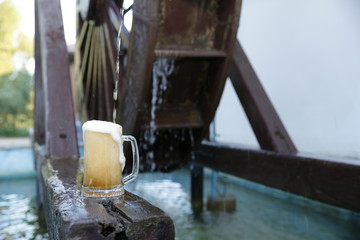 Glass of beer with foam on a natural wooden board on a background of a wooden mill wheel with falling water. Pattern, banner, background.
