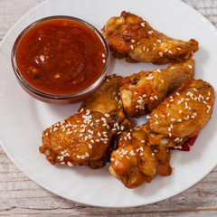 Fried juicy chicken wings marinated with honey, soy sauce, spices, sprinkled with sesame seeds on a white plate with sauce. Asian recipe, top view, close up