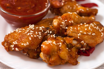 Fried juicy chicken wings marinated with honey, soy sauce, spices, sprinkled with sesame seeds on a white plate with sauce. Asian recipe, top view, close up