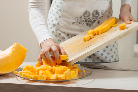 Kobieta w fartuchu kuchennym przekłada pokrojoną w kostkę dynie z drewnianej deski do krojenia na szklany talerz.