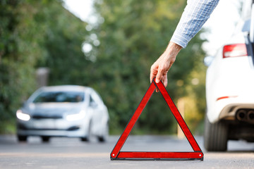 Close up of man driver putting red warning triangle/emergency stop sign behind his broken car on the side of the road, copy space and blurred car on background.