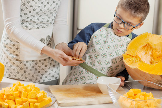 Kobieta i chłopiec w fartuchach kuchennych stoją przy blacie. Chłopiec zabiera z rąk kobiety świeże pestki dyni. Pokrojona w kostkę dynia leży na szklanym talerzu. 