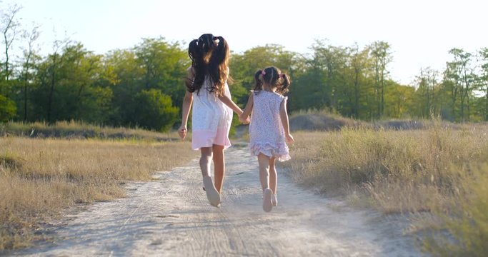 Back view whole plan two beautiful little girls of four and seven years old in summer dresses running dusty country  trail  in a meadow on warm summer evening. Slow motion 50 fps 4k