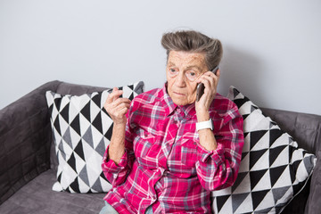 An old elderly woman grandmother with gray hair sits at home on the couch using the hand phone, a telephone conversation to hear the bad news. Emotion fear scare - obrazy, fototapety, plakaty