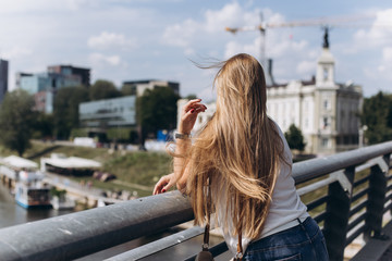 Fototapeta na wymiar Outdoors urban portrait of young hipster blondie woman in glasses with long hair smiling and sitting on bridge. Architecture, urban style, megalopolis concept