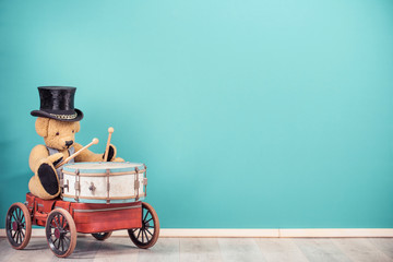 Retro Teddy Bear toy in old cylinder hat sitting on old luggage with wheels and playing the drum front mint blue wall background. Music concept. Vintage nostalgia style filtered photo