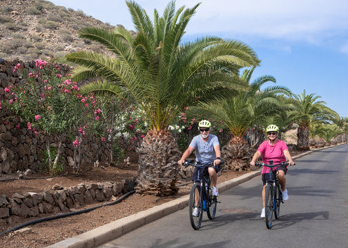 A cheerful senior couple man and woman enjoying freedom with electric bicycle. Happiness, fun and emotion outdoor in the street. Flowering plants and palm trees in background