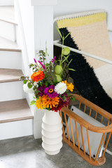Colorful orange, white, purple flower bouquet in vase next to chair and stairs, organic summer garden bouquet in home