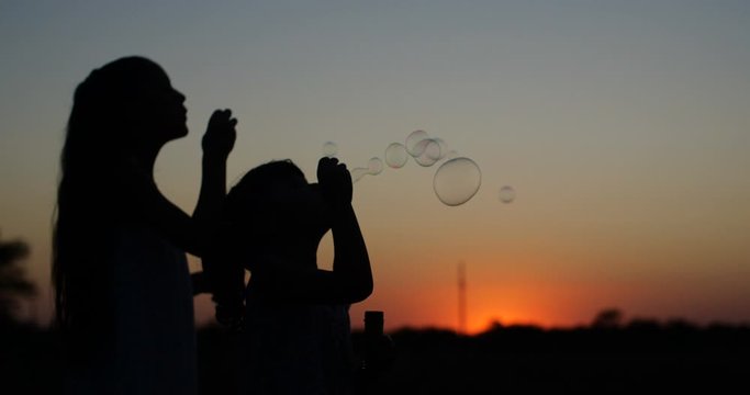 Silhouette of two pretty  girls of four and seven years old with long hair blowing  bubbles in the sunset of a warm summer day against a bright orange sky. 4k slow motion 50 fps