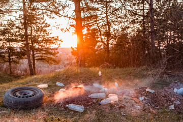 A car wheel next to a pile of burning junk in the forest.