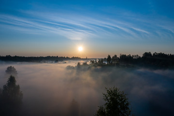 The rays of the morning sun make their way through the thick fog over the forest, field and river