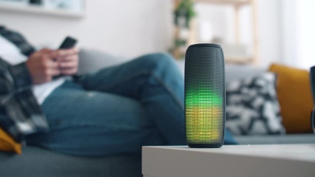 Man listens to music from a portable speaker that glows in different colors in the living room. 4k slow motion footage.