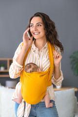 Young mother with little baby in sling talking by mobile phone at home