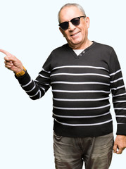 Handsome senior man wearing winter sweater and sunglasses with a big smile on face, pointing with hand and finger to the side looking at the camera.